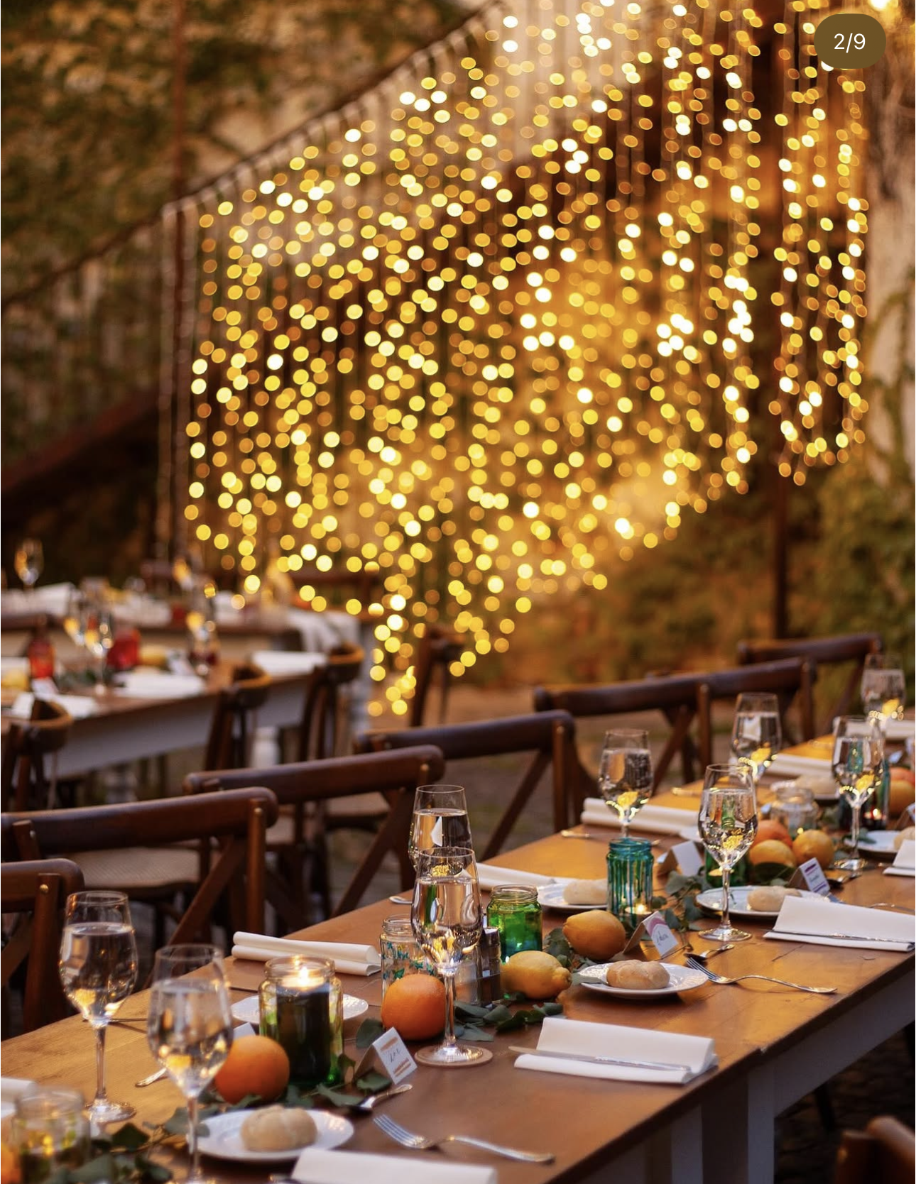 Curtain of warm fairy lights glowing above long candlelit dinner tables dressed with citrus and greenery